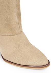 Botín de ante beige hasta el tobillo con punta afilada, diseño elegante y un sutil detalle de costura a lo largo del lateral, con suela de color claro.