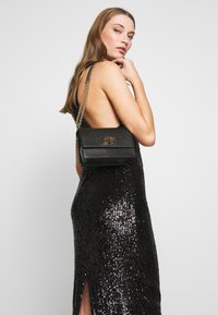 Borsa a tracolla in pelle nera con una catena, forma rettangolare, dettagli in metallo dorato e finitura testurizzata. Indossata sopra un abito con paillettes.