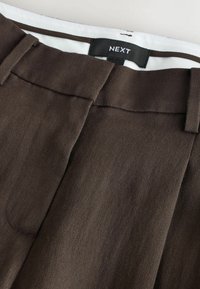 Pantalon marron sur mesure avec passants pour ceinture et une étiquette noire visible à la taille portant la mention « NEXT » sur une doublure blanche.