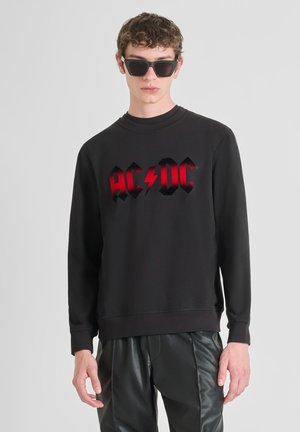 Sudadera negra con el logo rojo de "AC/DC" que presenta un rayo, escote redondo y puños de canalé, combinada con pantalones negros brillantes.