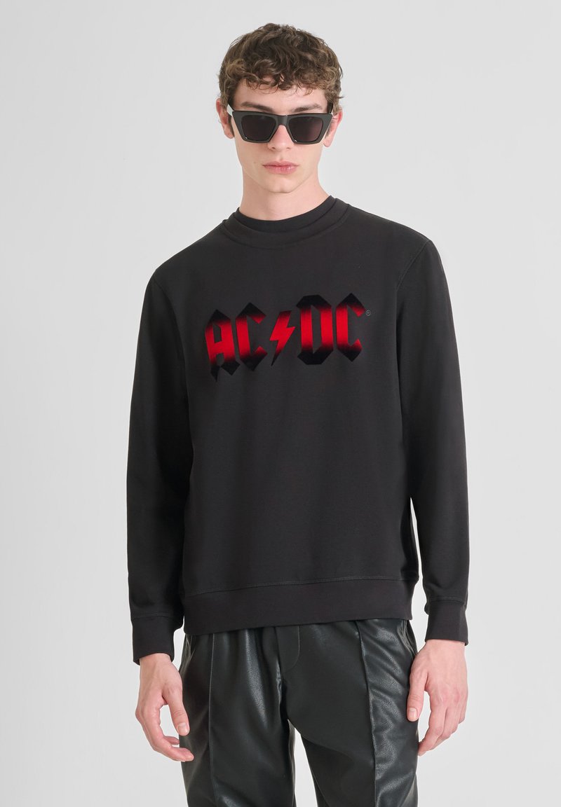 Zwarte sweatshirt met rood "AC/DC" logo met een bliksemschicht, ronde halslijn en geribbelde manchetten, gecombineerd met glanzende zwarte broeken.