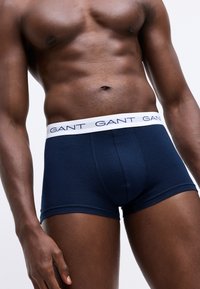 Torso maschile e parte superiore delle gambe che indossa boxer blu navy con vita bianca marchiata "GANT" su uno sfondo semplice.