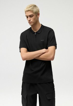 Młody mężczyzna z krótkimi blond włosami, ubrany w czarną krótko rękawową koszulkę polo i czarne spodnie typu cargo, stojący z założonymi ramionami na jednolitym tle.