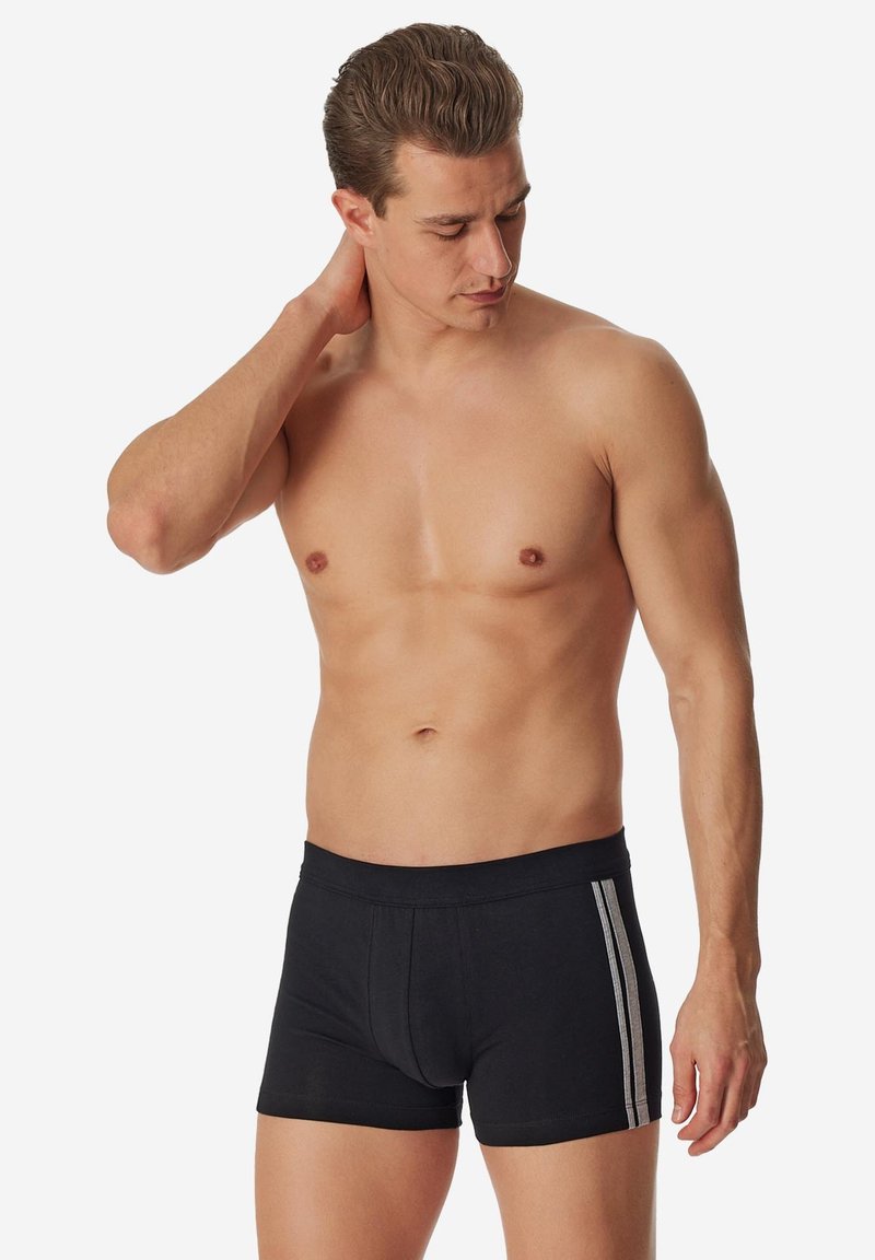 Uomo senza maglietta che indossa boxer neri con due strisce bianche ai lati, che guarda verso il basso toccandosi la nuca con una mano.