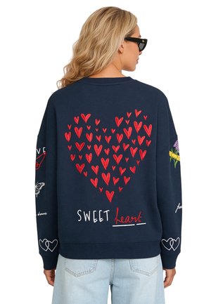 Femme aux longs cheveux blonds porte un sweat-shirt bleu foncé avec un motif de cœur rouge et le texte "Sweet heart" dans le dos, un jean bleu clair et des lunettes de soleil noires.