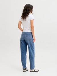 Γυναίκα με σγουρά σκούρα μαλλιά στέκεται, φορώντας λευκό μπλουζάκι, μπλε τζιν και λευκά αθλητικά παπούτσια, γυρισμένη με την πλάτη σε ένα απλό φωτεινό φόντο.