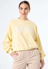 Mujer con cabello castaño que lleva pendientes de aro dorados, una sudadera amarilla con el texto "EL FUTURO ES AMOR" y pantalones de pana beige, con las manos en los bolsillos.