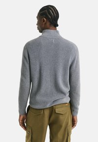 Mann mit geflochtenen Haaren, der einen grauen gerippten Rollkragenpullover und olivgrüne Cargo-Hosen trägt, von hinten vor einem schlichten Hintergrund gezeigt.