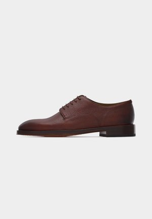 Zapato oxford clásico de hombre en cuero marrón con un tacón bajo y cierre de cordones cerrado, mostrado de perfil sobre un fondo claro y llano.