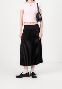 Kvinde iført en lyserød croppet t-shirt, sort plisseret midi-nederdel, hvidne strømper, sorte loafers og bærende en sort skuldertaske.