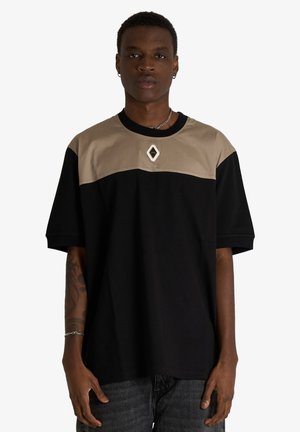 Giovane uomo che indossa una T-shirt con blocchi di colore nero e beige con un piccolo logo a forma di diamante sul petto, in piedi davanti a uno sfondo semplice.