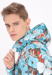 Jas met bloemenpatroon en capuchon in een blauwe, grijze en koraalkleurige kleurenschema. Gemaakt van lichtgewicht materiaal met zichtbare stiksels en ritsdetails.