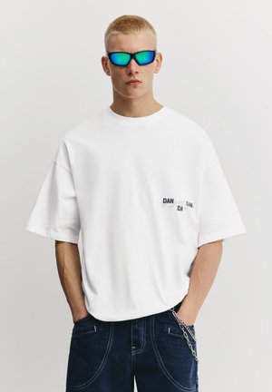 Maglietta bianca in cotone con vestibilità comoda e maniche corte; presenta una piccola grafica sul petto a sinistra. Indossata con occhiali da sole con lenti blu.