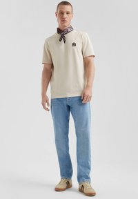 Polo shirt in cotone beige con una piccola patch del logo, abbinato a jeans azzurri chiari e sneakers beige con strisce nere. Sciarpa stampata attorno al collo.