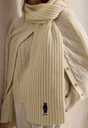 Femme portant un pull en maille torsadée crème, une écharpe assortie avec un petit ours brodé et un pantalon blanc, les mains dans les poches, sur un fond neutre.