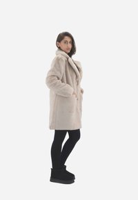 Beige faux fur frakke med bred krave, sidelommer og en afslappet pasform. Båret med sorte leggings og ankelstøvler. Glat tekstur.
