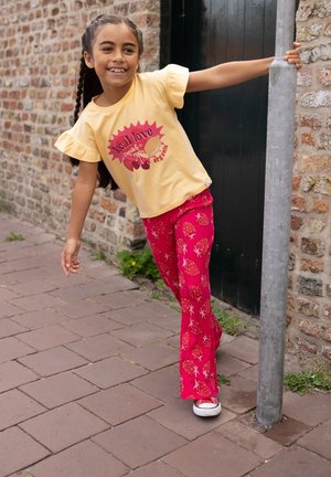 Jong meisje dat glimlacht en leunt op een paal, draagt een geel t-shirt en rode broek met aardbeienpatroon op een stenen straat.