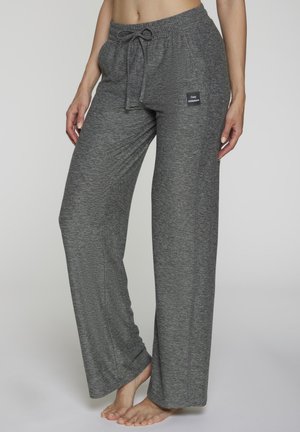 Woman wearing loose gray drawstring lounge pants with "Cozy Athleisure" patch, standing barefoot against a plain background.