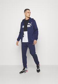 Námořnická mikina s bílým logem Puma, kombinovaná s černobílým grafickým tričkem, ladícími námořnickými tepláky a černými sportovními botami.