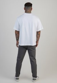 Camiseta blanca de manga corta con un ajuste relajado, combinada con pantalones grises y zapatos blancos. El fondo es liso y de un gris claro.
