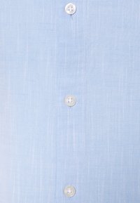 Camicia di lino azzurro chiaro con tessuto texture, caratterizzata da sottili linee verticali e sei bottoni bianchi sul davanti.