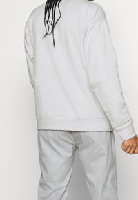 Persona con el cabello trenzado que lleva una sudadera gris claro y pantalones gris claro, de pie contra un fondo blanco liso.