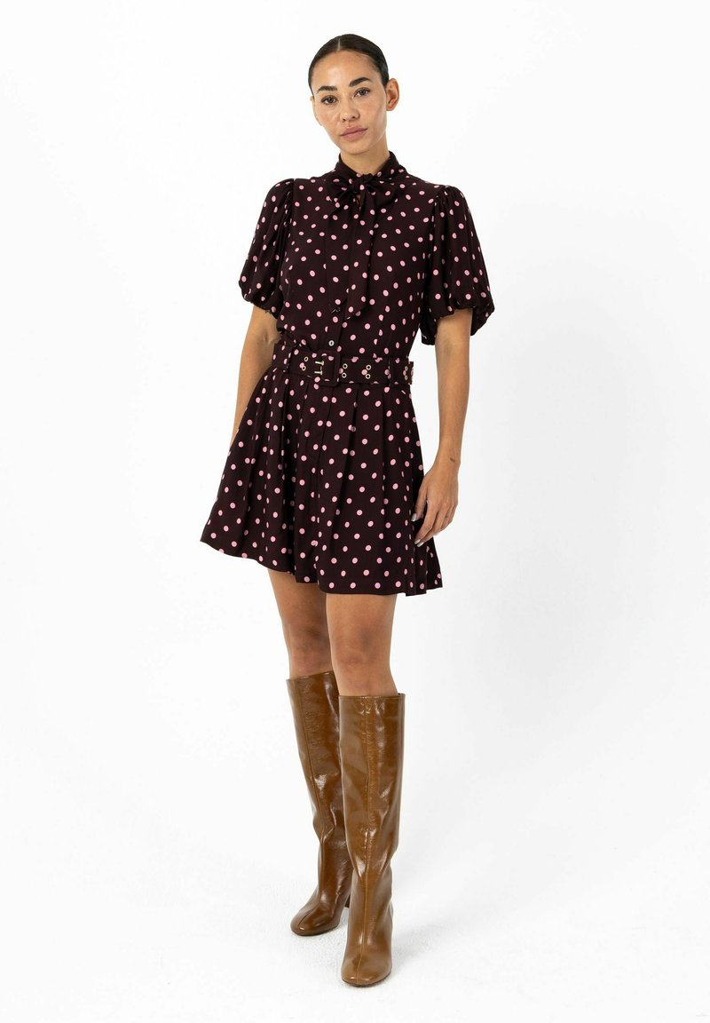 Robe bordeaux foncé à pois roses, manches courtes bouffantes, boutonnée sur le devant, taille ceinturée et jupe évasée. Bottes marron montant jusqu'aux genoux.