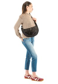 Bolso negro con lentejuelas brillantes en el frente, forma curva, correa ajustable, llevado cruzado. Modelo con suéter beige y jeans azules.