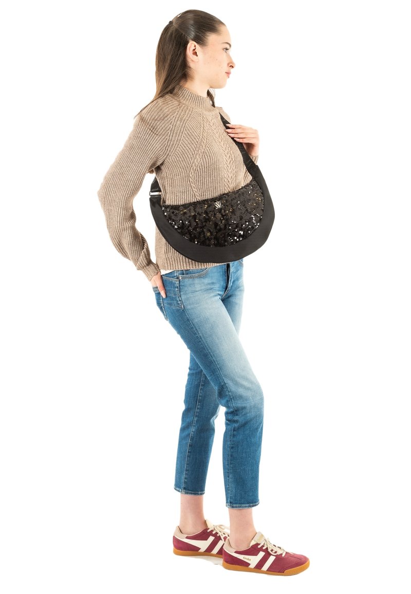 Bolso negro con lentejuelas brillantes en el frente, forma curva, correa ajustable, llevado cruzado. Modelo con suéter beige y jeans azules.
