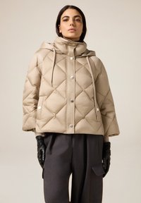 Beige doorgestikte gewatteerde jas met capuchon, voorzien van een knoopsluiting aan de voorkant, zilverkleurige knopen en een trekkoorddetail. Draagt donkergrijze broek.