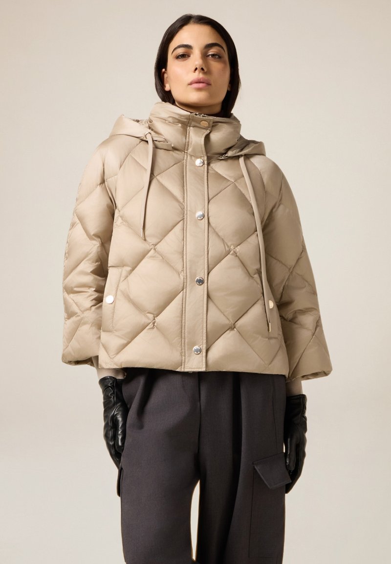 Beige doorgestikte gewatteerde jas met capuchon, voorzien van een knoopsluiting aan de voorkant, zilverkleurige knopen en een trekkoorddetail. Draagt donkergrijze broek.