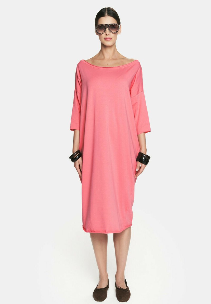 Fuksjowa, oversize sukienka z szerokim dekoltem i rękawami o długości trzech kwartałów. Gładki materiał opada do połowy łydki. Para czarnych bransolet.