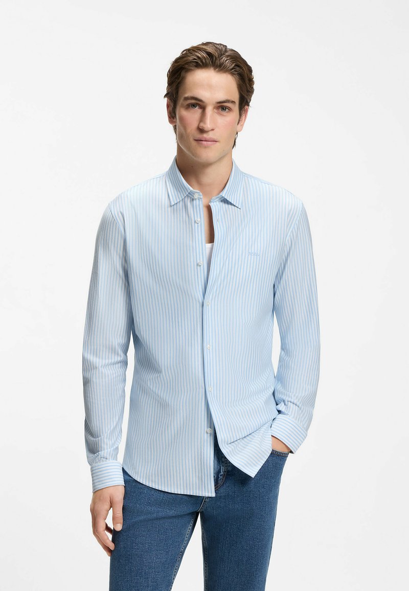 Jeune homme portant une chemise à rayures bleu clair et blanc par-dessus un sous-vêtement blanc et un jean bleu, debout avec une main dans la poche.