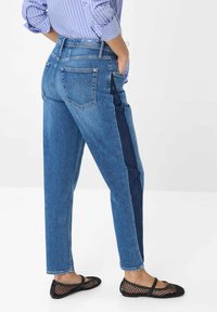 Persona che indossa jeans in denim blu bicolore con una striscia laterale scura, una camicia a righe blu e scarpe piatte in mesh nere, in piedi con le mani in tasca.