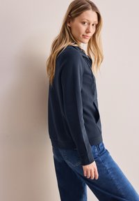 Hoodie blu navy con tasca frontale, maniche lunghe e chiusura a mezza zip. Tessuto morbido, vestibilità comoda. Abbinato a jeans blu. Sfondo chiaro.