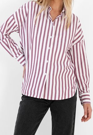 Woman wearing a white and mauve vertical striped button-up shirt with black pants, standing with one hand on her hip.