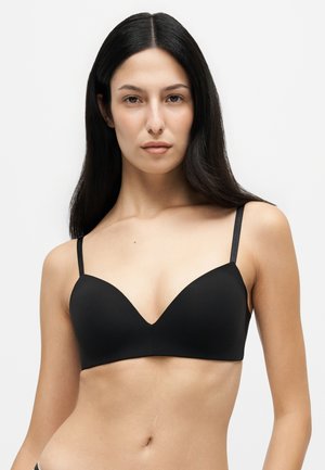 Femme aux longs cheveux foncés portant un soutien-gorge noir à fines bretelles, face à un fond blanc uni.