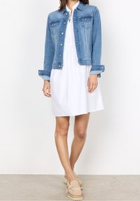 Vaaleansininen farkkutakki, jossa on napit edessä, rintataskut ja käännetyt hihat, yhdistettynä valkoiseen mekkoon ja vaaleanbeigeihin korokepolkimiin.