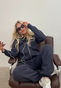Marine blå jakke med glidelås laget av myk stoff, parret med hvite sneakers. Modellen sitter på en brun lærstol, iført solbriller.