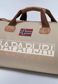 Borsa duffle beige con dettagli in pelle marrone, dotata di un patch con la bandiera norvegese e di un logo bianco in evidenza con testo arancione. Manico testurizzato.