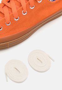 Oranžová semišová teniska s bílými tkaničkami a kovovými průchodkami, vybavená strukturovanou gumovou podrážkou a kulatou špičkou.