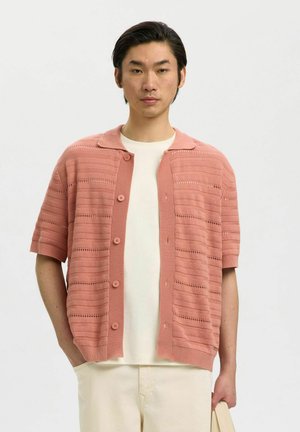 Mann kledd i ferskenfarget kortermet knappet cardigan over hvit t-skjorte og beige bukser, står med en hånd i lommen og holder en bag.