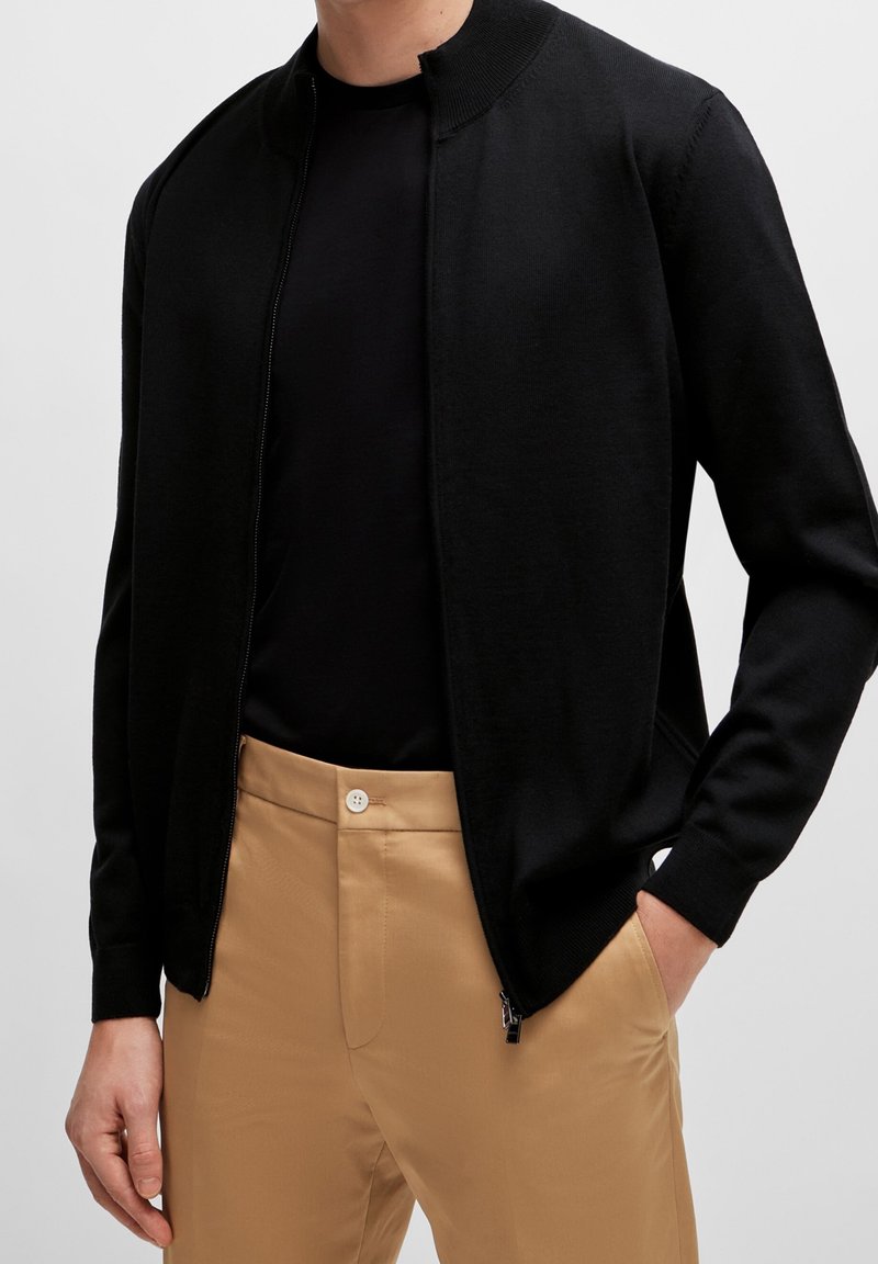 Homme portant un cardigan noir zippé par-dessus une chemise noire et un pantalon beige, une main dans la poche, sur un fond gris uni.