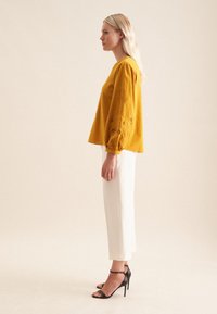 Blusa gialla senape con dettagli ricamati sulle maniche, abbinata a pantaloni crema e sandali neri con tacco alto.