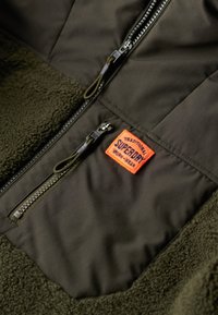 Giacca in pile e tessuto verde con tasca con zip, toppa logo arancione ed elementi testurizzati. Il design include zip resistenti.