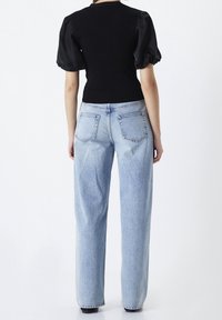 Femme portant un haut noir à manches bouffantes et un jean taille haute bleu clair à jambes droites, debout de dos devant un fond blanc.