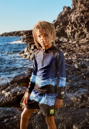 Blauwe rash guard met golvenpatroon en korte mouwen, gecombineerd met zwarte zwembroek met een logo en felgroene accenten, bij een rotsachtig strand.