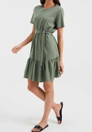 Femme portant une robe verte à manches courtes avec un lien à la taille et un ourlet volanté, marchant pieds nus dans des sandales noires à enfiler avec une boucle dorée.