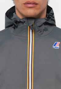 Chaqueta con capucha gris y cierre en el frente. Presenta un patrón a rayas en amarillo, rojo y azul. Incluye el logo de K-Way en el pecho.