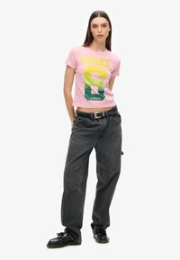 Rosa, kurz geschnittenes T-Shirt mit „OSAKA 6“-Grafik in Gelb und Grün, kombiniert mit locker sitzenden, dunkelgrauen Jeans und schwarzen Schnürschuhen.